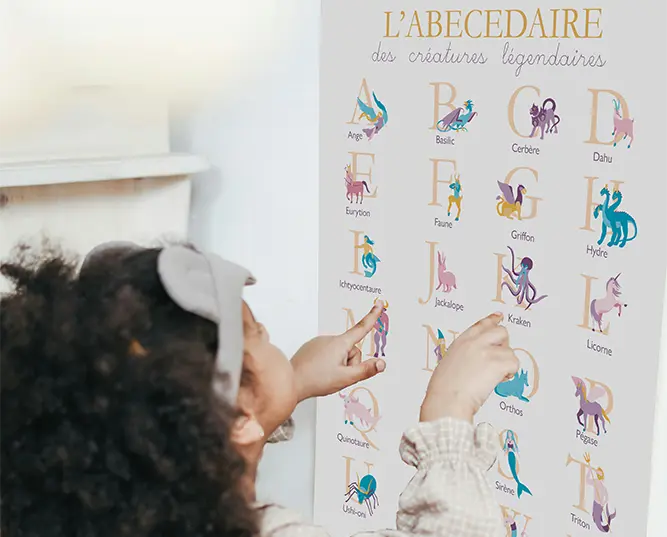 Petite fille interagissant avec l'affiche "L'abécédaire des créatures légendaires", montrant des lettres et des illustrations fantastiques, Yblou Création.